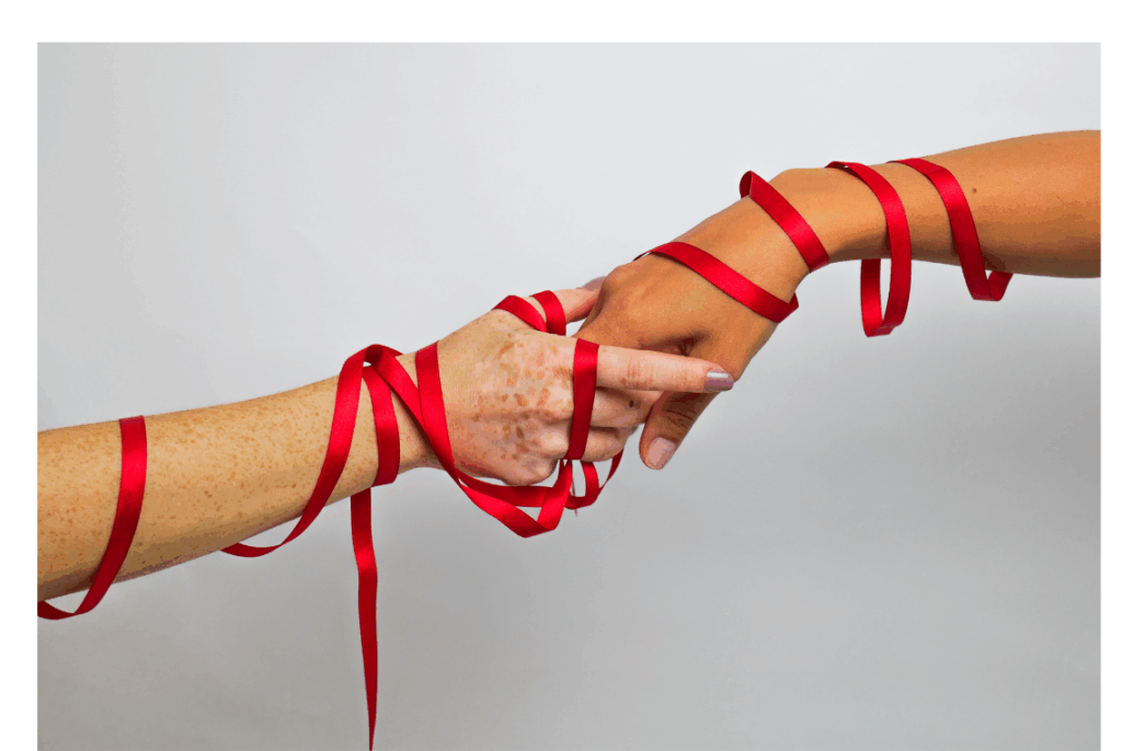 Two hands of different skin tones wrapped together with a red ribbon, symbolizing connection, unity, or support.