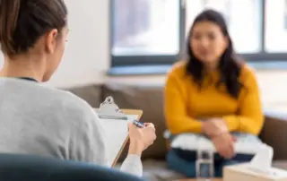 A female counsellor holding a clipboard and pen listens attentively to a woman in a yellow sweater during a therapy session in a well-lit room.