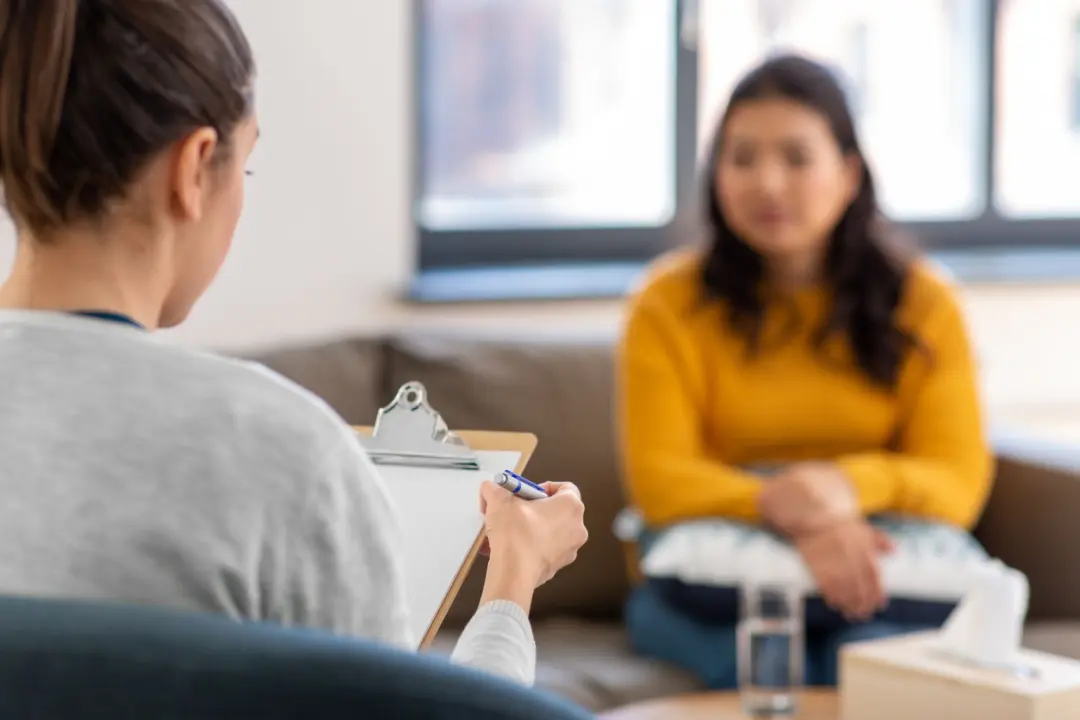 A female counsellor holding a clipboard and pen listens attentively to a woman in a yellow sweater during a therapy session in a well-lit room.