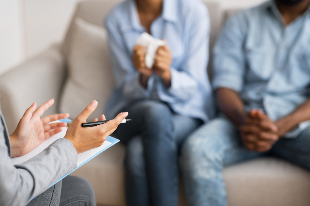A counsellor gestures while holding a clipboard and pen, facing a family members seated on a couch. One person is holding tissues, suggesting emotional distress.