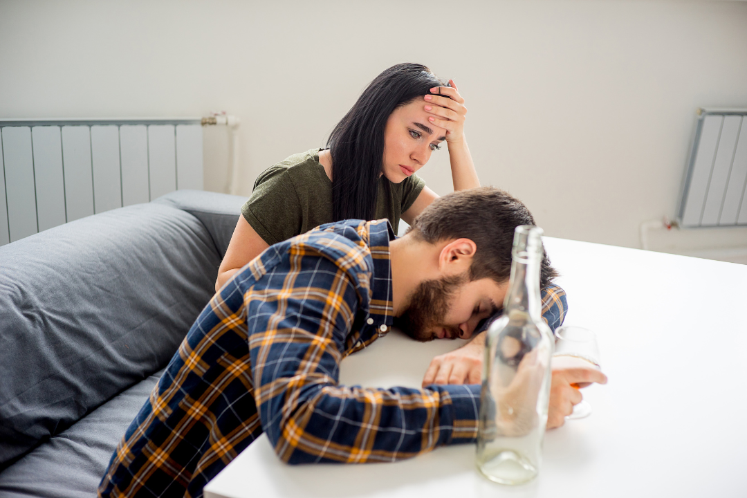 Worried woman comforting a distressed man with alcohol in front of him, highlighting the emotional toll of supporting a partner struggling with substance use.