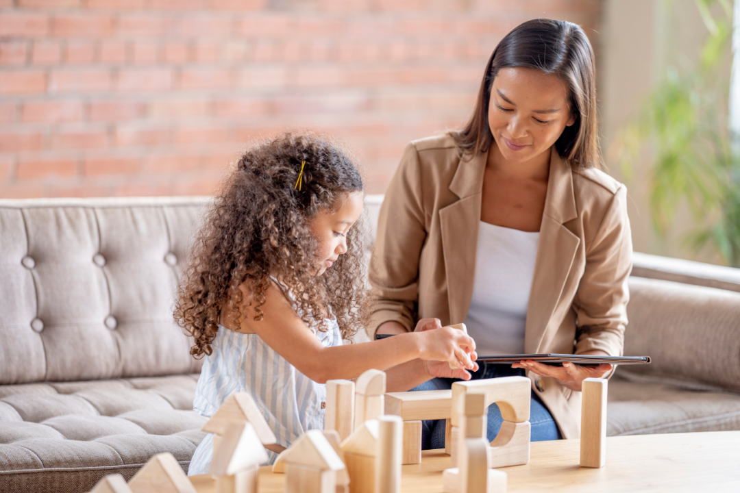 Child building with wooden blocks while a therapist observes at eye level in a calm, home-like therapy room.