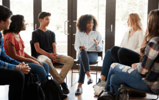 A female counsellor leads a relaxed group discussion with diverse young people seated in a circle in a bright room, symbolising support and supervision.