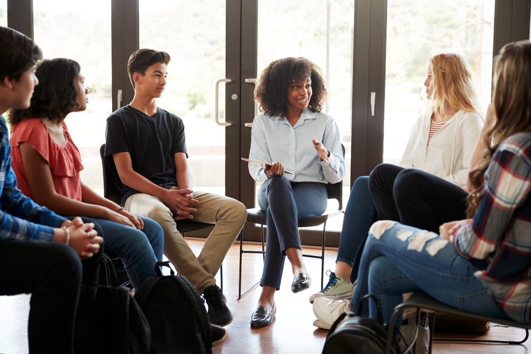 A female counsellor leads a relaxed group discussion with diverse young people seated in a circle in a bright room, symbolising support and supervision.