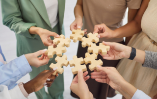 A group of people stand together holding wooden puzzle pieces in a circle, symbolising teamwork, collaboration, connection, and shared purpose.