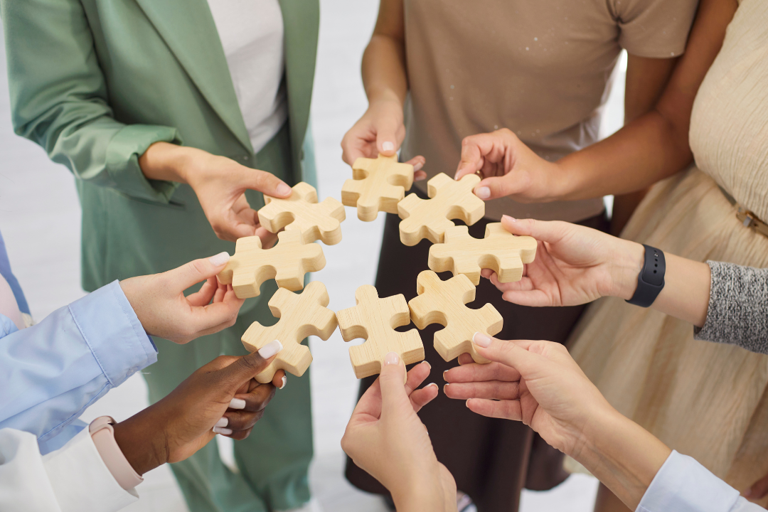 A group of people stand together holding wooden puzzle pieces in a circle, symbolising teamwork, collaboration, connection, and shared purpose.